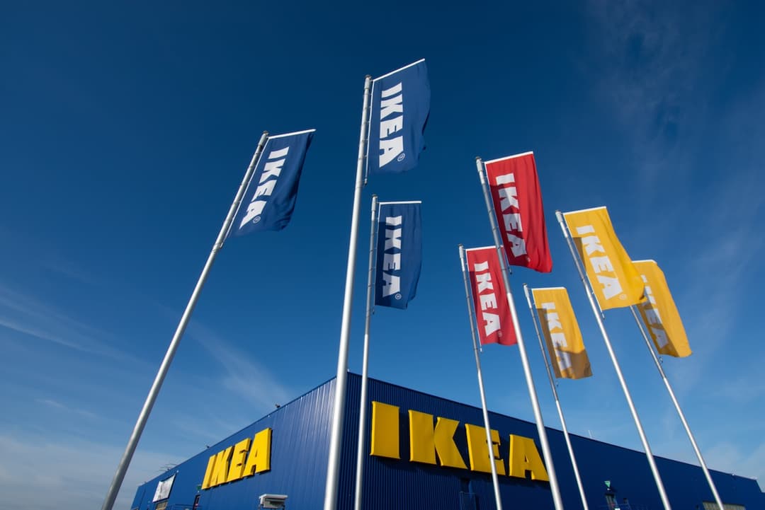 Banderas de IKEA ondeando en lo alto de mástiles frente a una tienda IKEA de color azul con letras amarillas, bajo un cielo azul claro