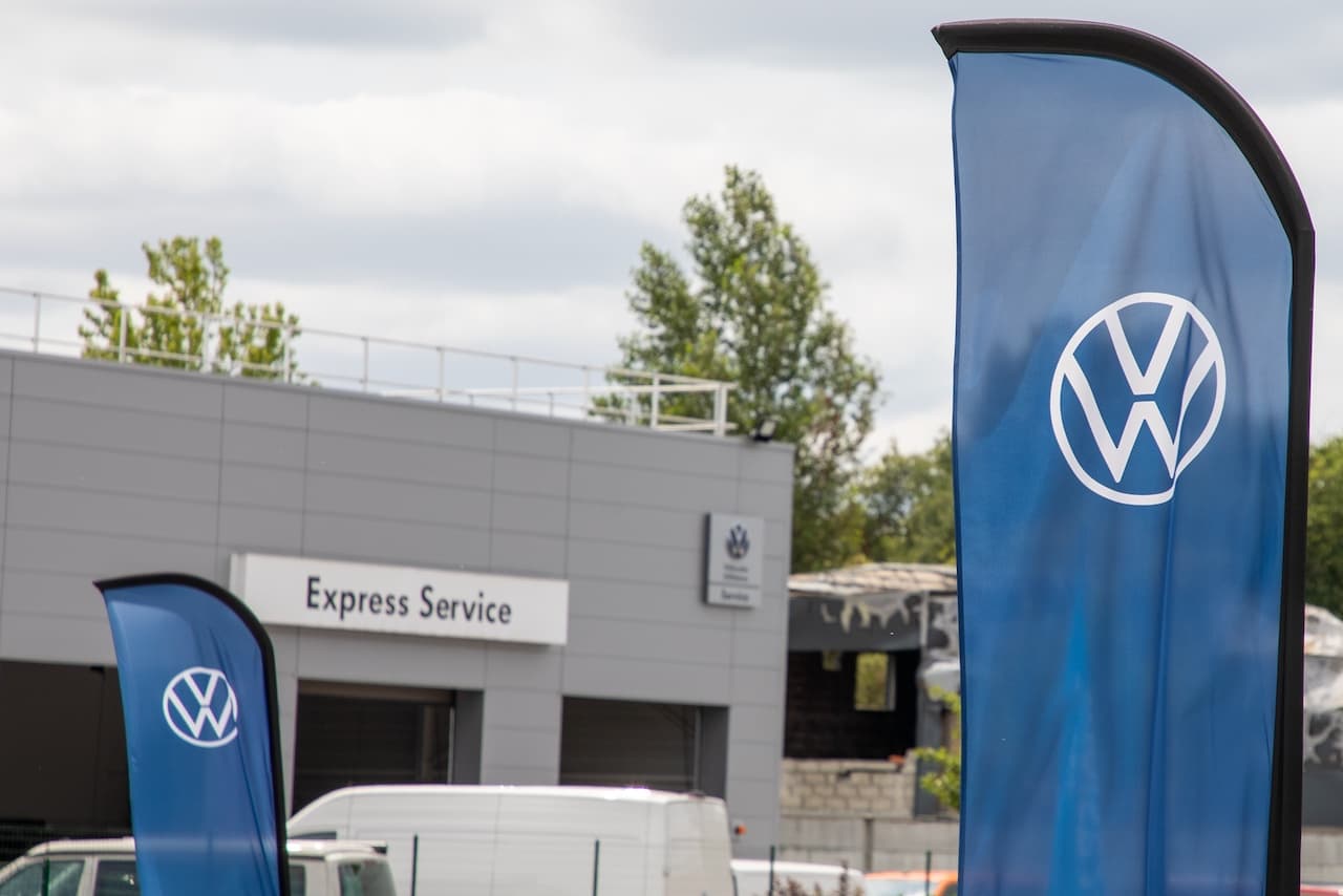 Se observan banderas azules con el logotipo de Volkswagen en primer plano, y al fondo se distingue un edificio que indica 'Express Service', también de Volkswagen. Hay vegetación alrededor y un vehículo blanco estacionado.