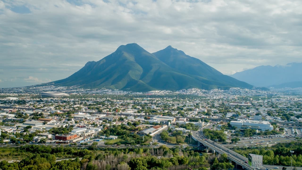 Una vista panorámica de Monterrey, donde se destacan las montañas del cerro de la Silla al fondo. Se observa una extensa área urbana con edificios y calles, así como un estadio visible en el centro. La imagen refleja la combinación de naturaleza y desarrollo urbano, subrayando el paisaje característico de la región, un punto clave del norte de México.