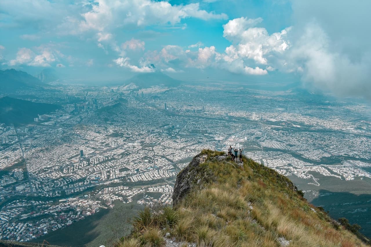 Un grupo de personas se encuentra en la cima de una montaña, contemplando un vasto paisaje urbano con edificios y áreas verdes debajo. El cielo está parcialmente nublado, con tonos azules y nubes blancas. El lugar parece ofrecer una vista panorámica impresionante de la ciudad. El entorno natural se combina con la urbanización, destacando la conexión entre ambos. Este escenario puede representar una experiencia única de aventura y exploración, un 'punto clave' de conexión entre la naturaleza y la vida urbana.