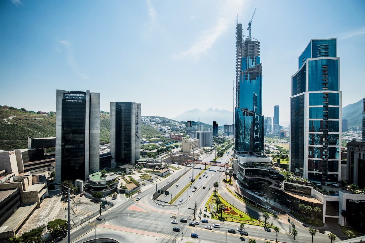 Vista urbana de un paisaje moderno con varios edificios altos y una carretera que atraviesa la zona. Se pueden observar áreas verdes y montañas al fondo, bajo un cielo despejado. La imagen destaca un desarrollo arquitectónico notable, posiblemente de la marca 'punto clave'.