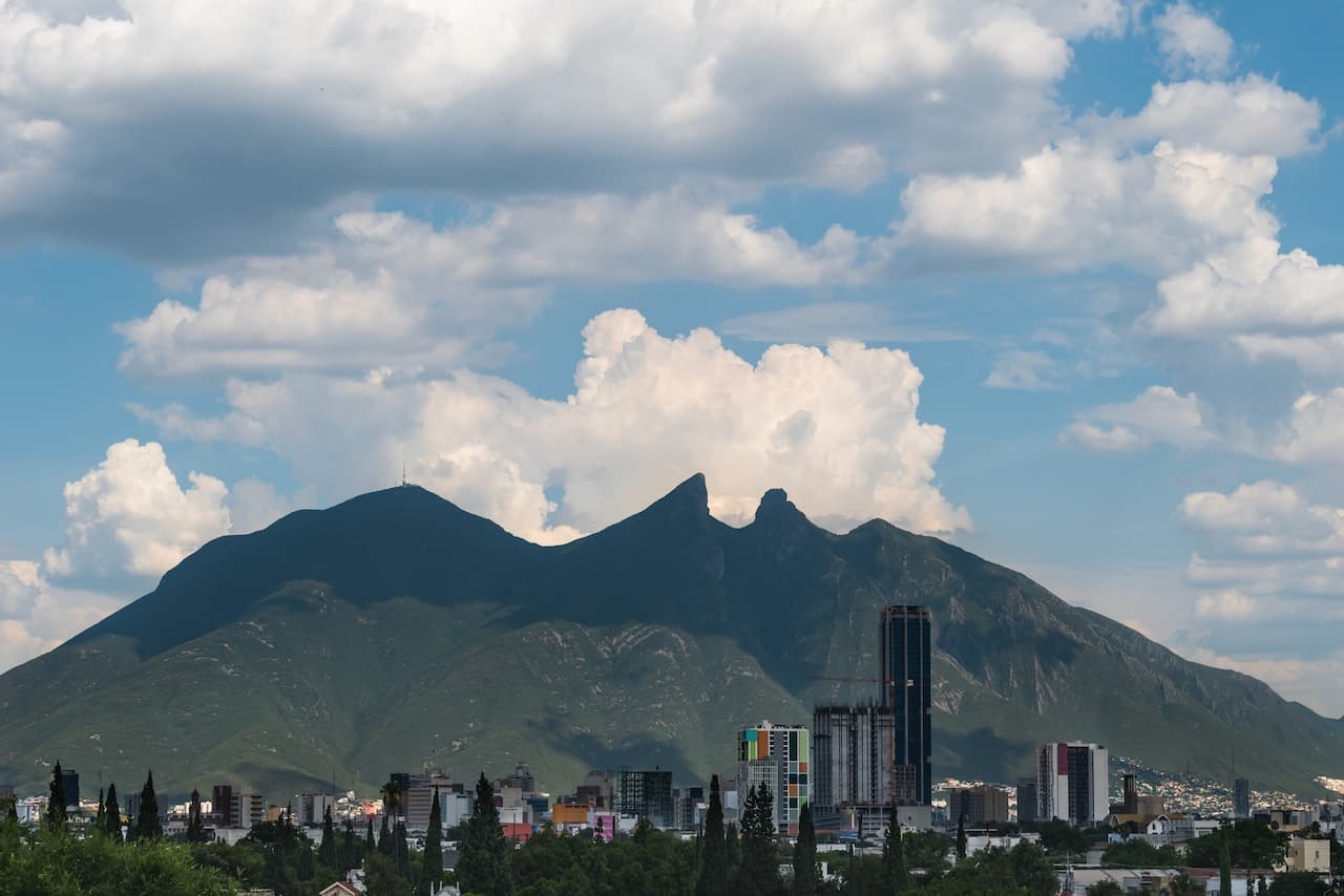 Vista panorámica de montañas con nubes y una ciudad en primer plano. Las montañas tienen una forma distintiva y están rodeadas de edificios y árboles. El cielo es mayormente azul con nubes blancas, destacando el paisaje natural y urbano. Esta escena es característica de la región de Monterrey, destacando la marca 'punto clave' del lugar.