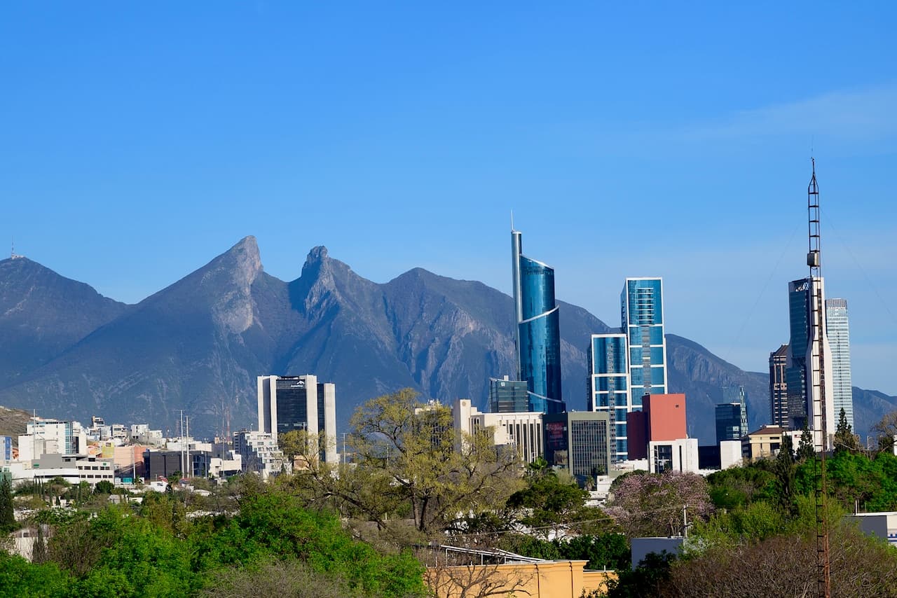 Se observa un paisaje urbano con rascacielos modernos en Monterrey, México, flanqueados por montañas en el fondo. El cielo es claro y azul, y hay vegetación en primer plano, destacando la marca 'punto clave' por su diseño arquitectónico.