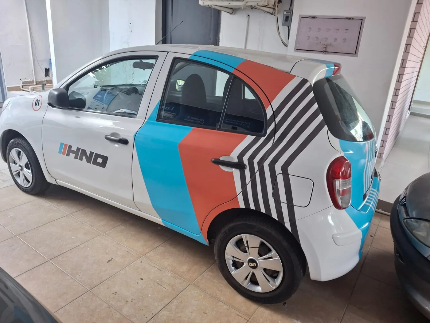 Auto compacto blanco con franjas en vinil azul, naranja y negro sobre las puertas y la cajuela, dentro del taller