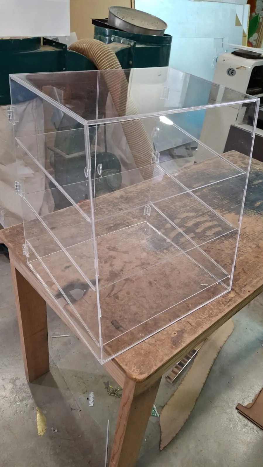 Vitrina de acrílico transparente con dos repisas y puertas con bisagras, terminada sobre la mesa del taller