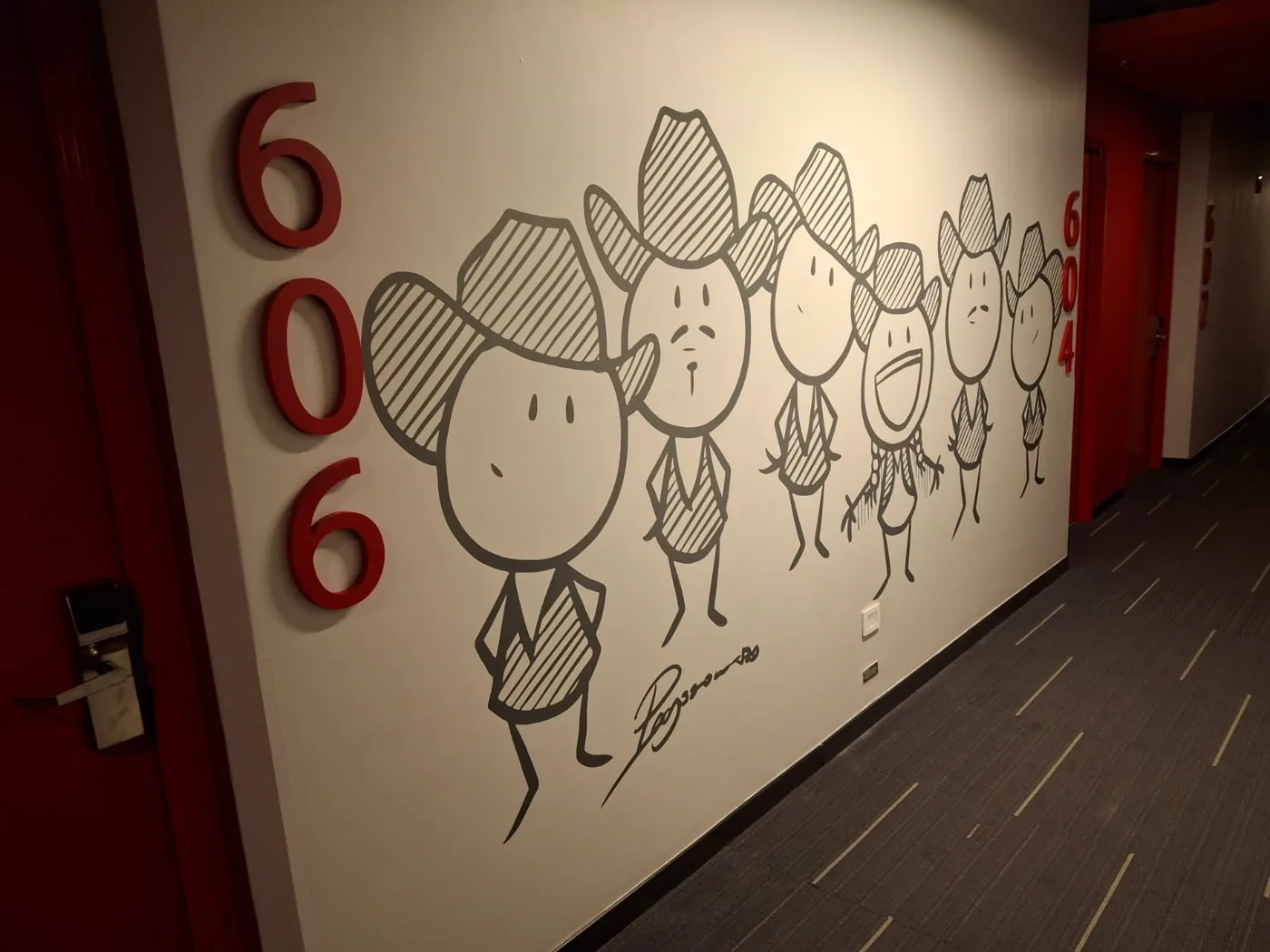Muro de pasillo con personajes de sombrero dibujados en vinil gris junto a un número de habitación en rojo