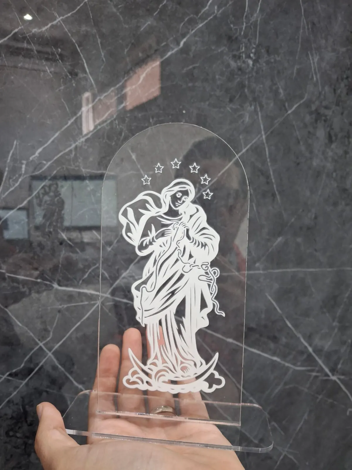 Figura religiosa grabada sobre acrílico transparente cortado en arco con base, sostenida en la palma de la mano