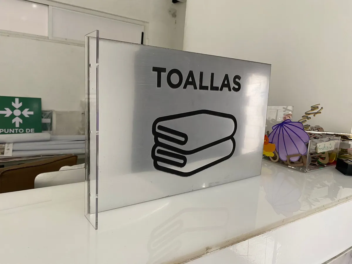 Placa plateada con la palabra Toallas y un ícono negro, montada en un soporte de acrílico transparente