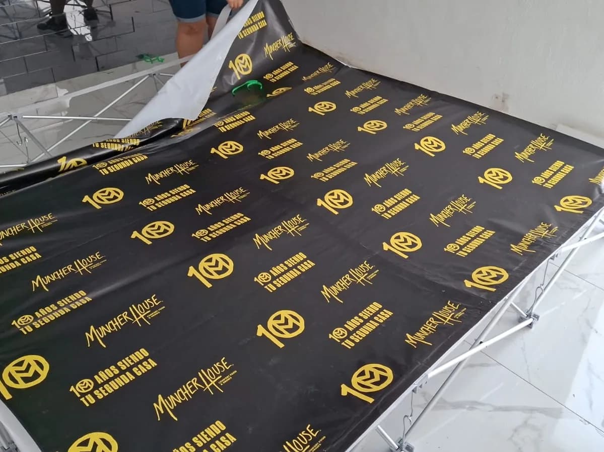 Lona negra con logotipos dorados repetidos, mientras se tensa sobre un bastidor plegable para backdrop de evento