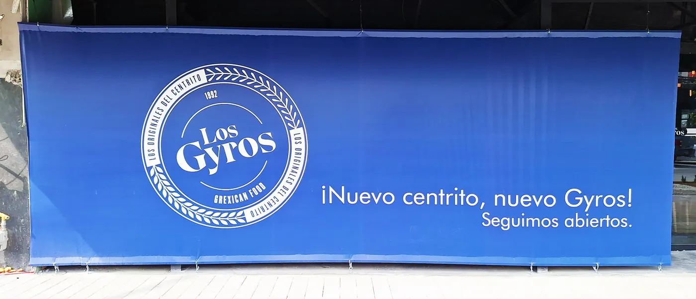 Lona azul de gran formato con ojillos, fijada a una reja frente a un local en remodelación