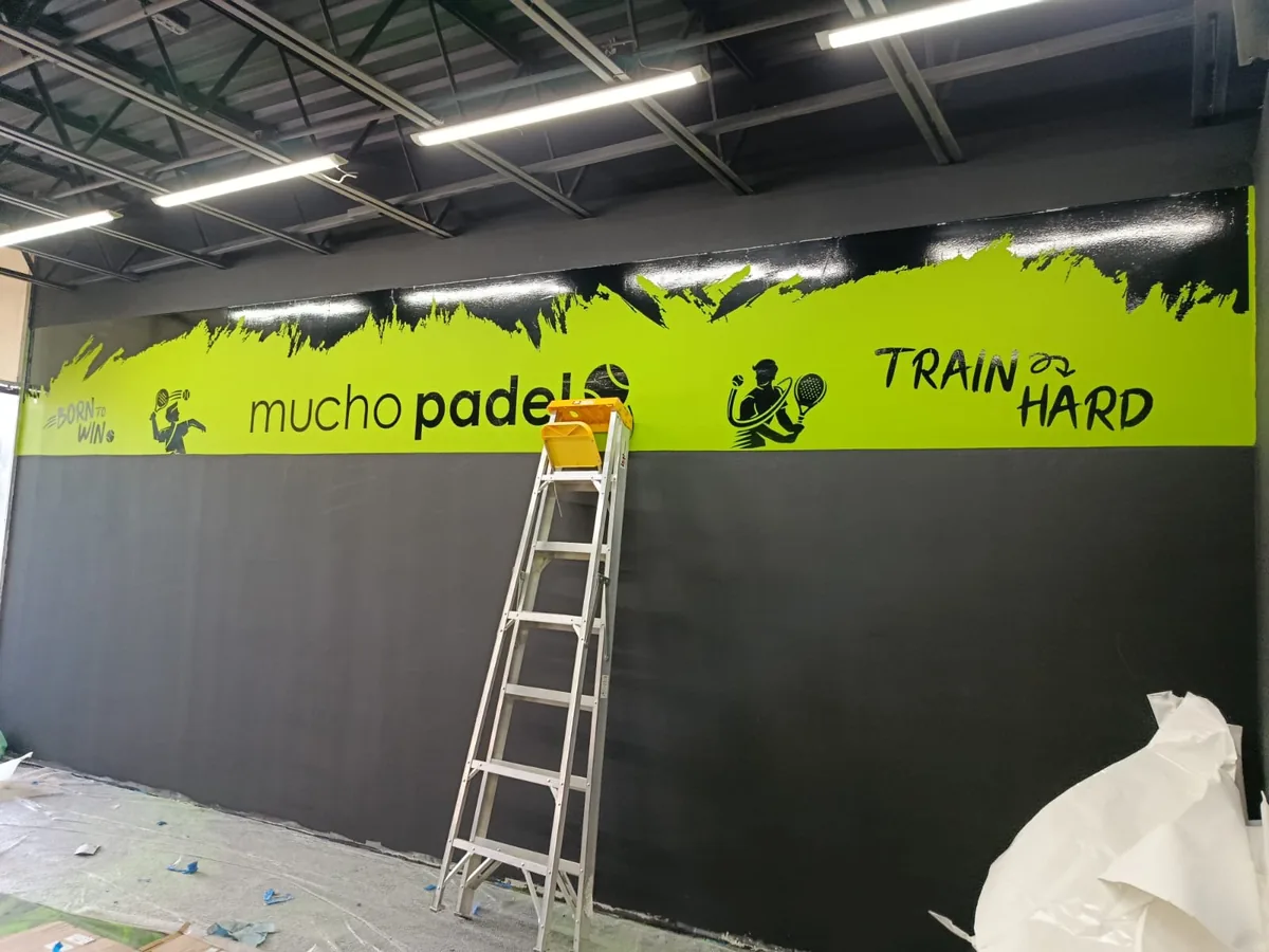 Muro de un club de pádel con franja verde, letras y siluetas de jugadores en vinil, con la escalera del instalador al frente